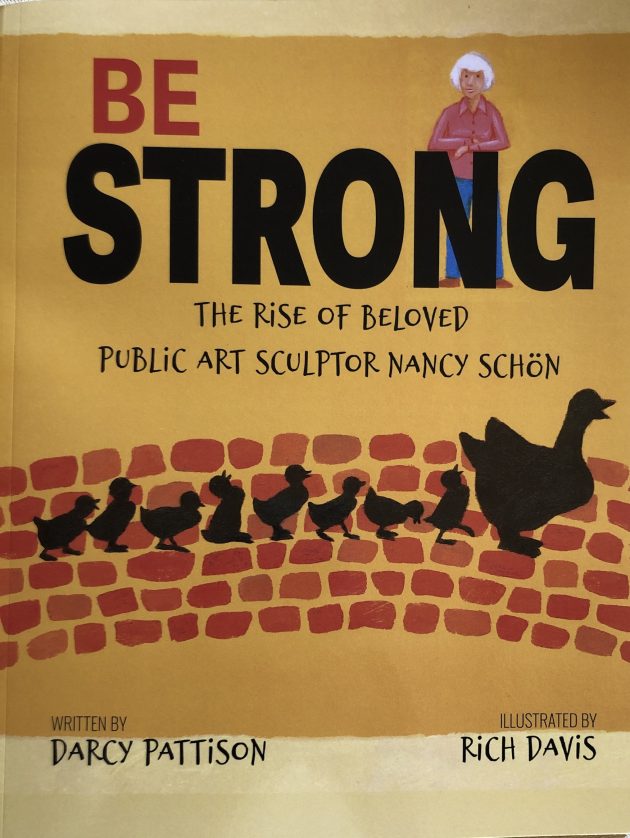 Cover art showing sculptor Nancy Schön, an older woman with white hair, leaning on the letter "N" in the book title BE STRONG: THE RISE OF BELOVED PUBLIC ART SCULPTOR NANCY SCHÖN. A mother duck and her eight ducklings walk along a brick pathway under the title